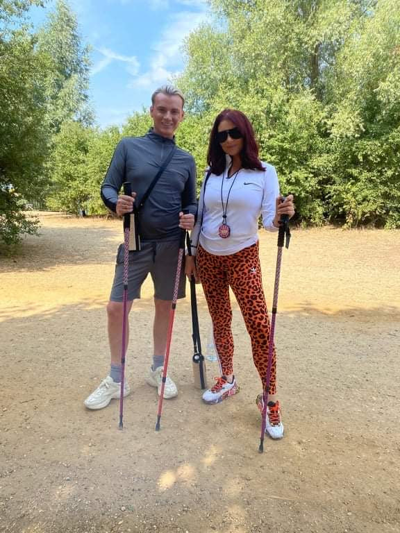 Two people standing outdoors with hiking sticks, one wearing a grey outfit and the other in a white top and red patterned leggings.