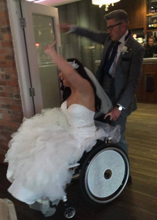 A lady wearing a wedding dress in a wheelchair being pushed by a man in a grey suit 